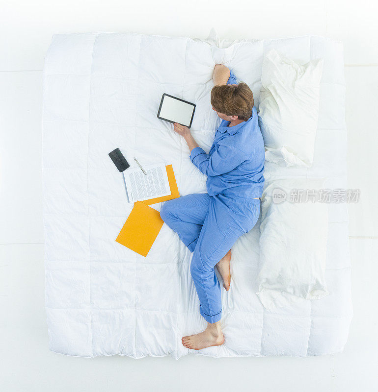 白人年轻男子/男性穿着睡衣在卧室的白色背景前睡觉/放松/休息，使用智能手机/数码平板电脑